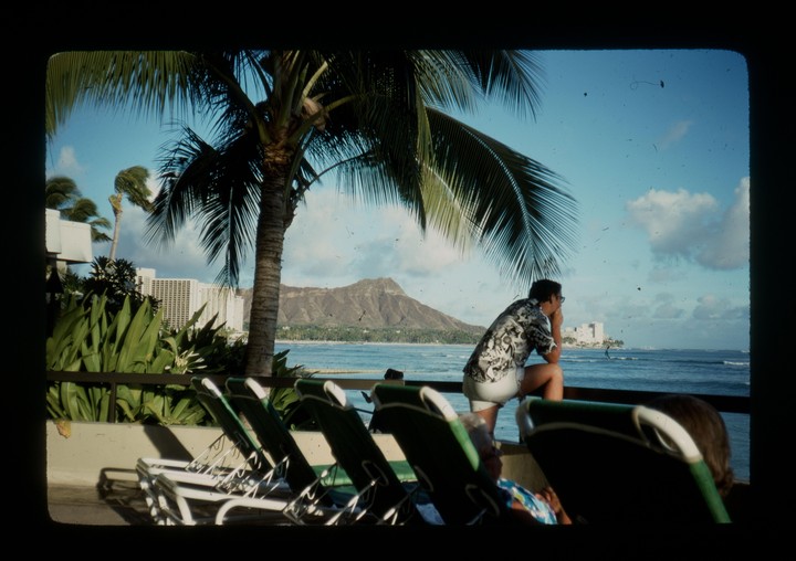 A man in a patterned shirt and shorts leans on a railing, looking out at the ocean with a mountain in the distance, framed by palm trees and lounge chairs.