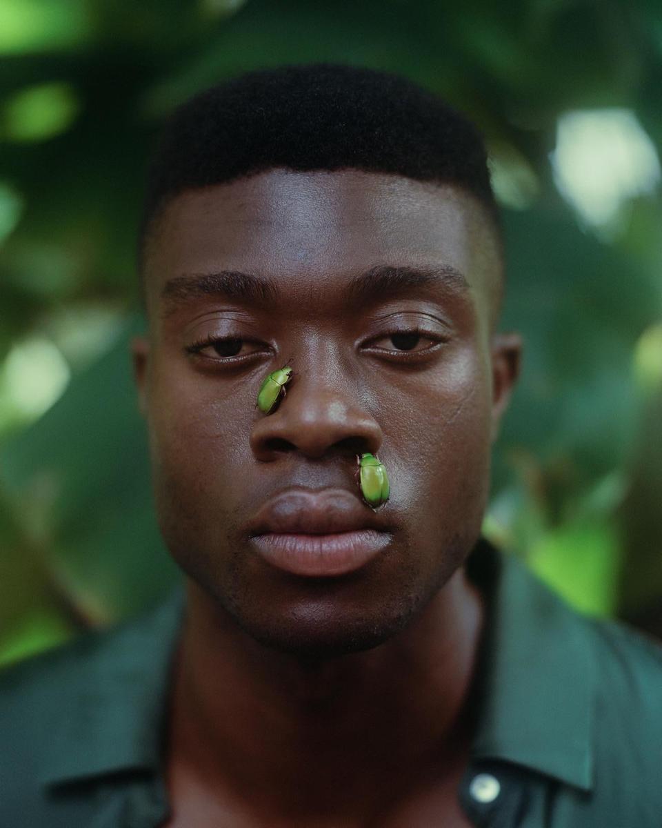A man with two green beetles on his face looks directly at the viewer.