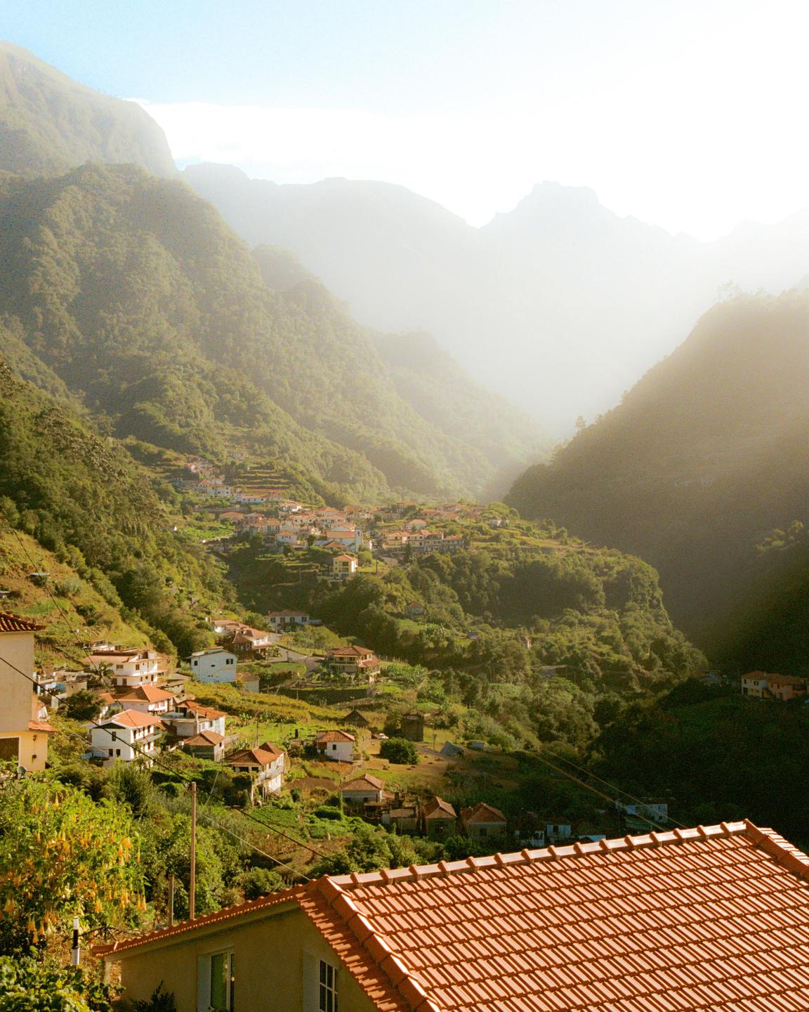 A village with many houses and red tile roofs is nestled in a green valley between mountains.