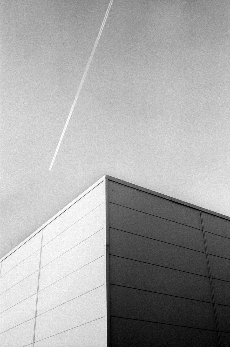 A contrail streaks across a bright sky above a building with two distinct sides, one light and one dark.