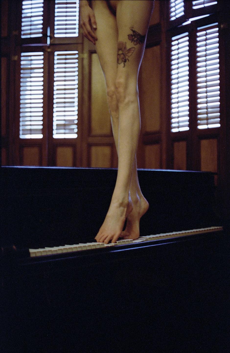 A woman's legs and bare feet are visible as she stands on the keys of a piano, with light coming through window blinds in the background.