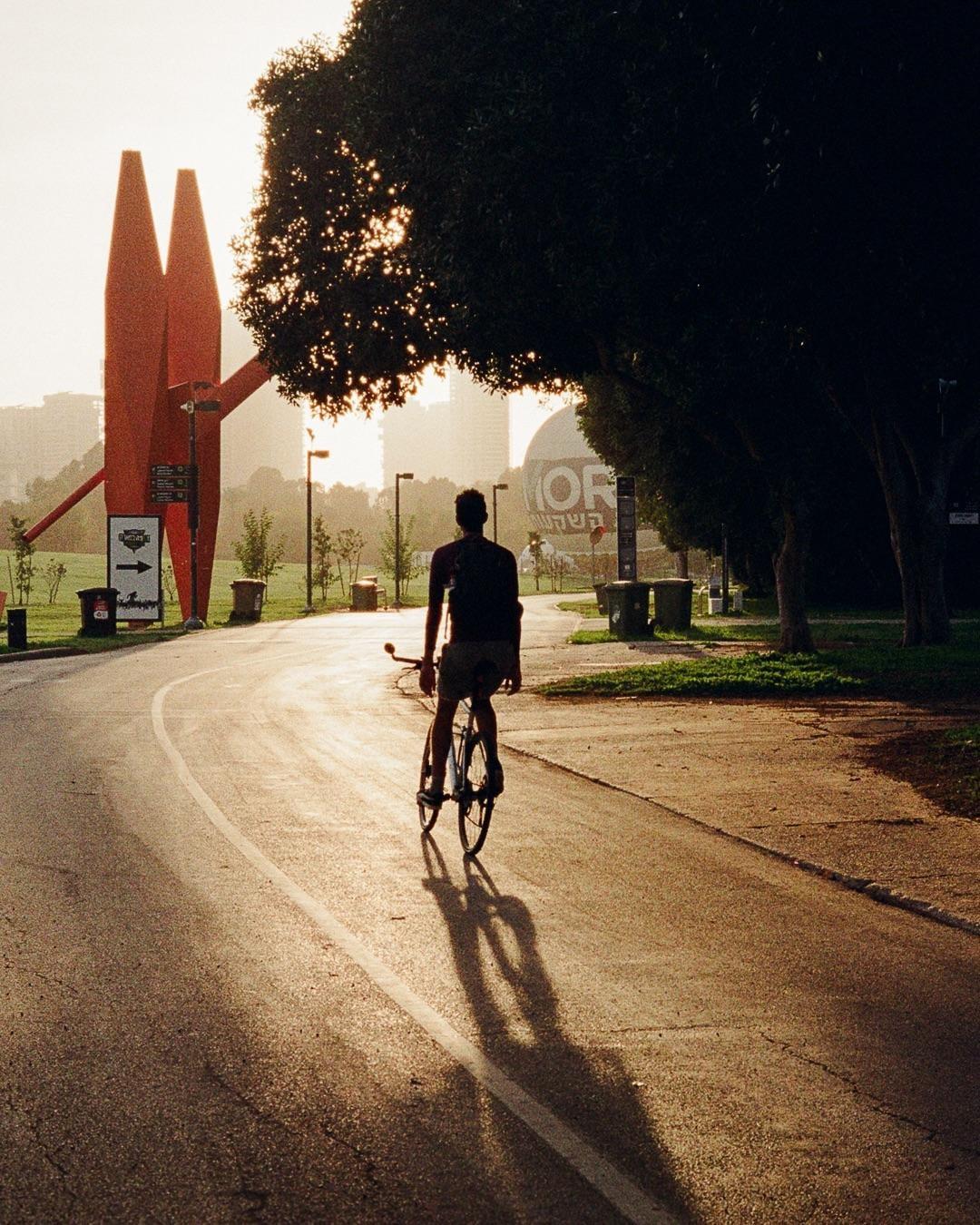 A man rides a bicycle on a paved path with a large red sculpture and trees in the background.