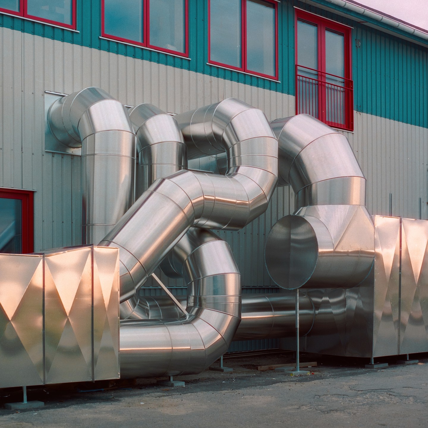 Large silver ventilation pipes are attached to the side of a building with red-framed windows and a teal and gray facade.