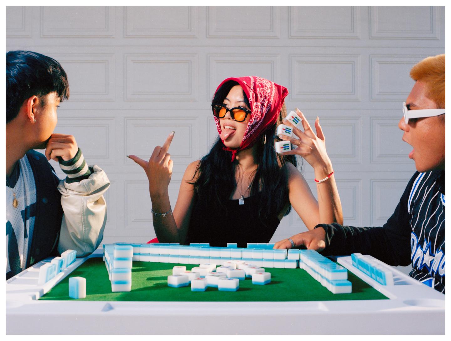 Three people are playing mahjong at a table with a white garage door in the background.