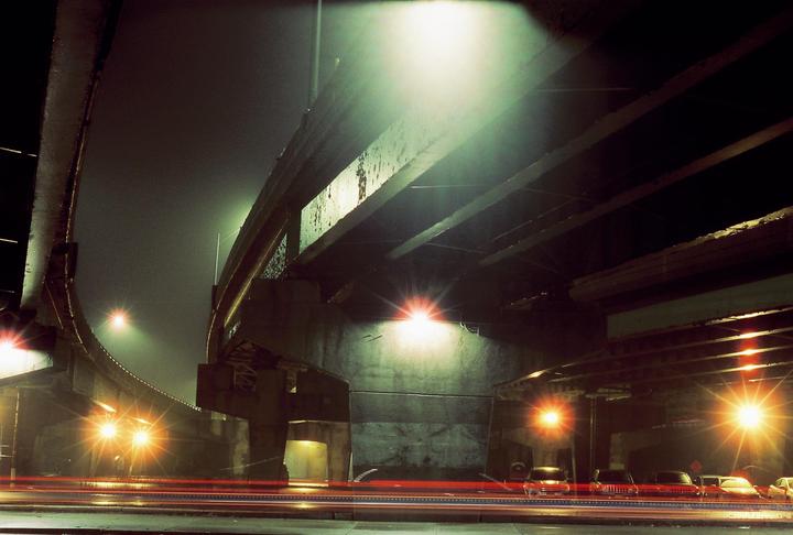 A long exposure shot captures the light trails of vehicles under a highway overpass at night.