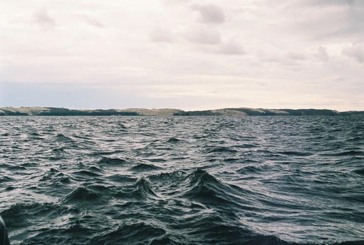 A body of water with waves is in the foreground, and a distant landmass with dunes and trees is under a cloudy sky.