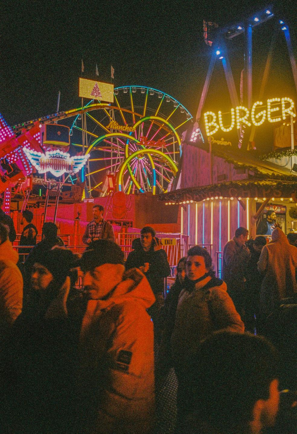 A crowd of people stands in front of a brightly lit Ferris wheel and a "BURGER" sign at night.