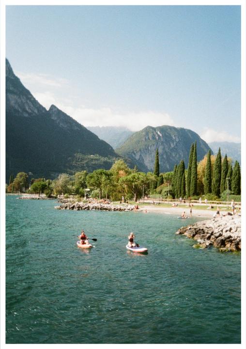 Two people paddleboard on a lake with mountains and a beach in the background.