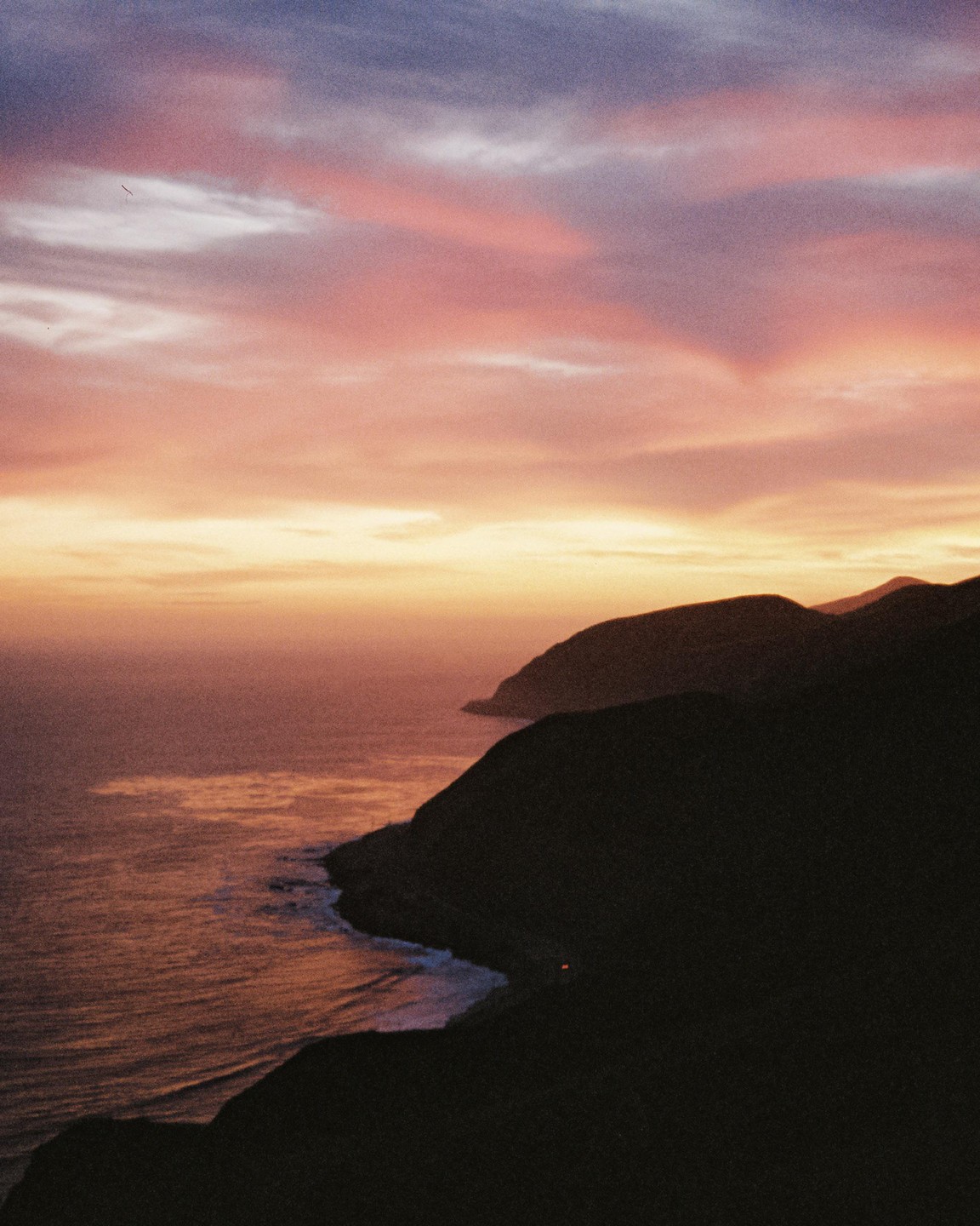 The ocean meets a dark coastline under a colorful sunset sky.