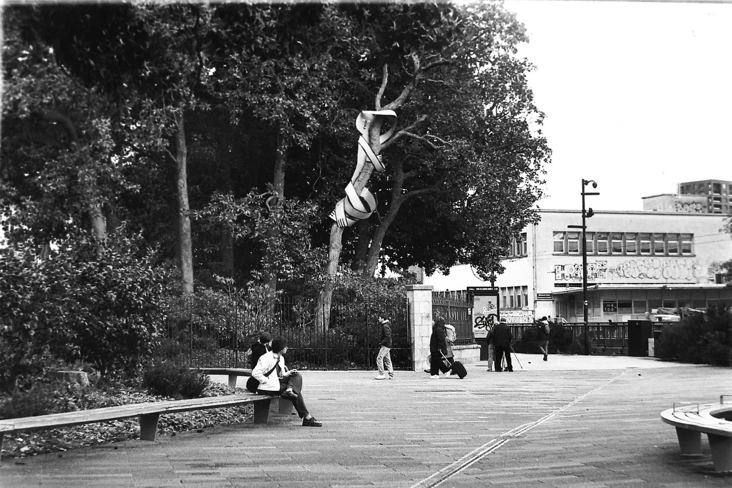 A black and white street scene shows people walking and sitting on a bench, with a tree wrapped in fabric in the background.