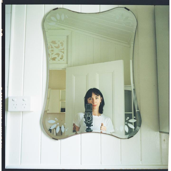 A woman with dark hair holds a camera and looks into a decorative mirror, capturing her reflection.