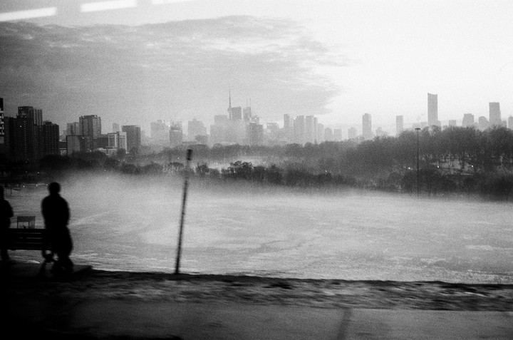 A hazy, black and white cityscape of Toronto is seen from a distance, with a person sitting on a bench in the foreground.