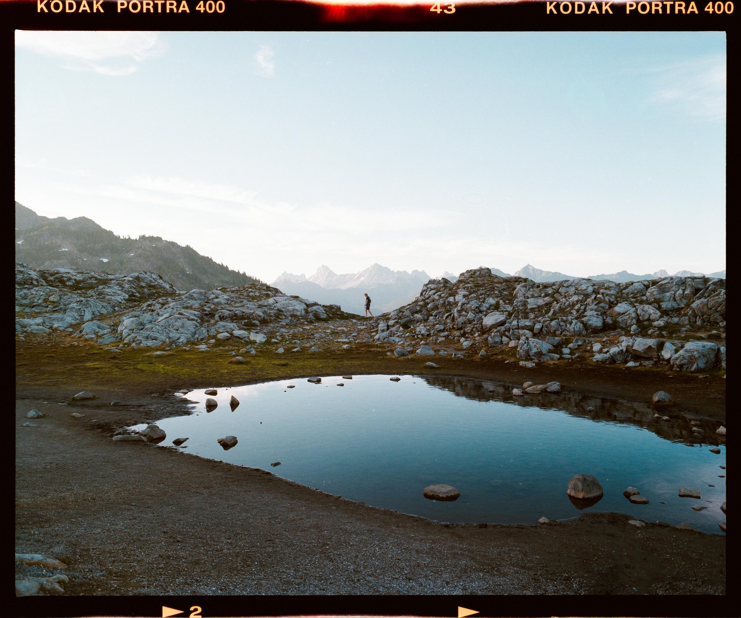 A person walks along a rocky path next to a small pond with mountains in the background.
