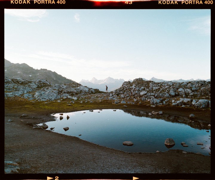 A person walks along a rocky path next to a small pond with mountains in the background.