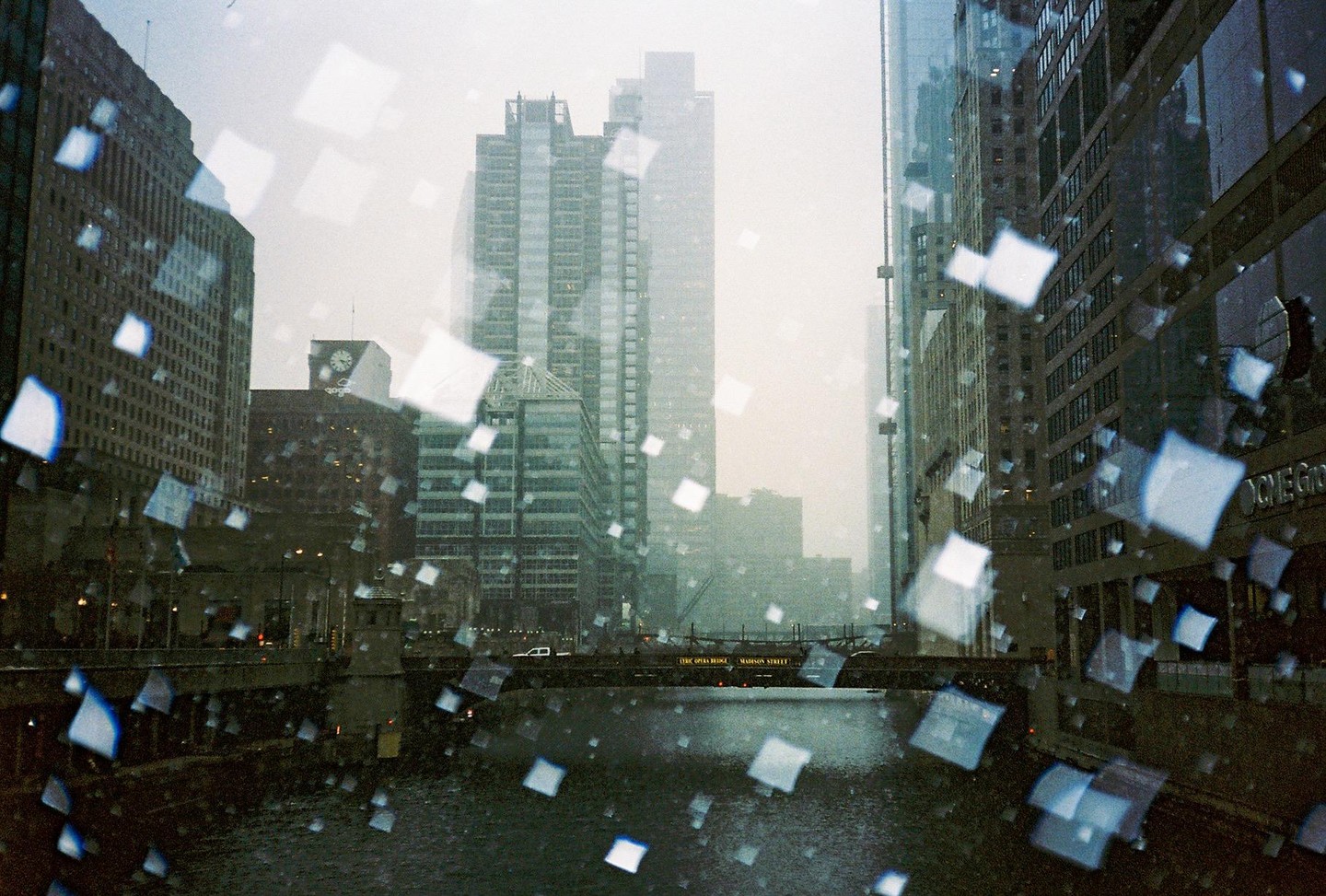 A city street is seen through a window covered in square-shaped water droplets, with a river and bridge in the foreground and tall buildings in the background.