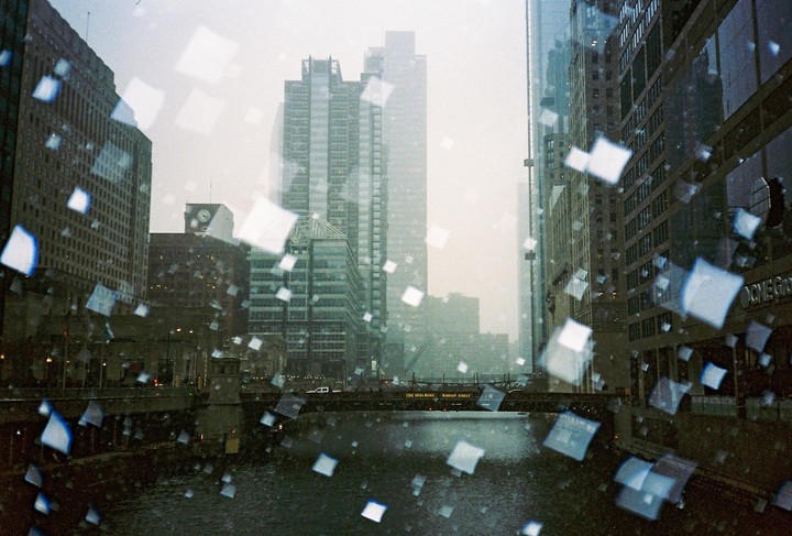 A city street is seen through a window covered in square-shaped water droplets, with a river and bridge in the foreground and tall buildings in the background.