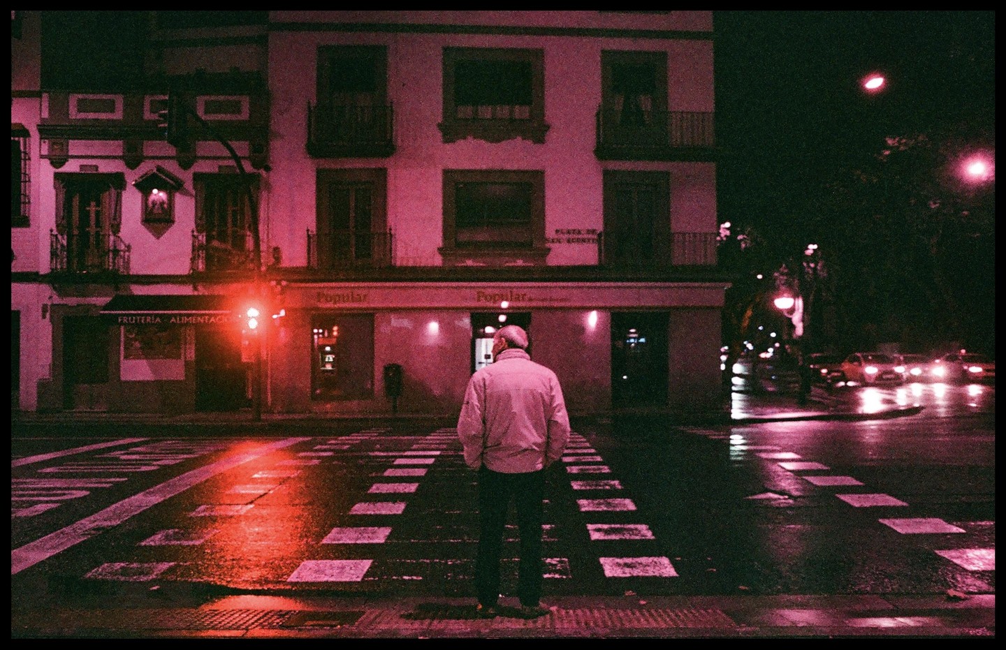 A man stands on a wet crosswalk at night, facing a building with shops on the ground floor.