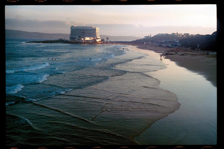 Waves roll onto a beach with a large hotel and a town on a hill in the background at sunset.