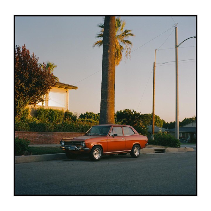 An orange car is parked on a residential street next to a palm tree at sunset.