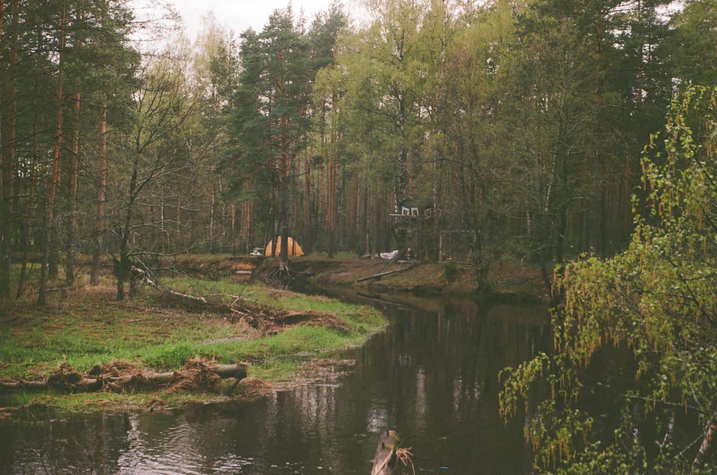 A river flows through a forest with a tent and a treehouse visible on the bank.