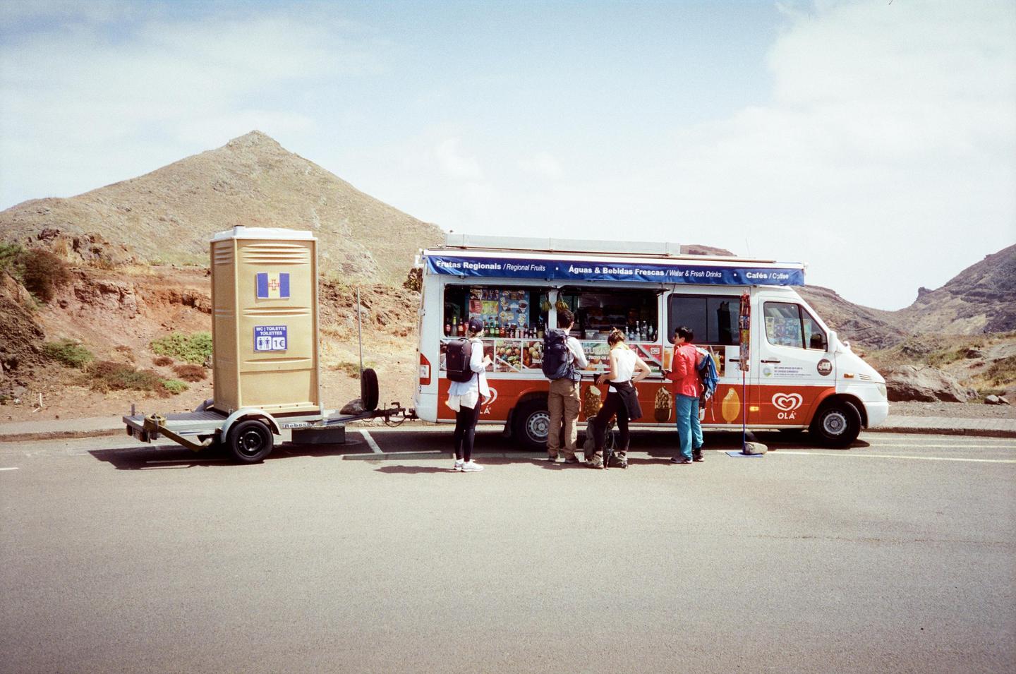 A group of people stand around an ice cream truck with a portable toilet on a trailer next to it, in a mountainous landscape.