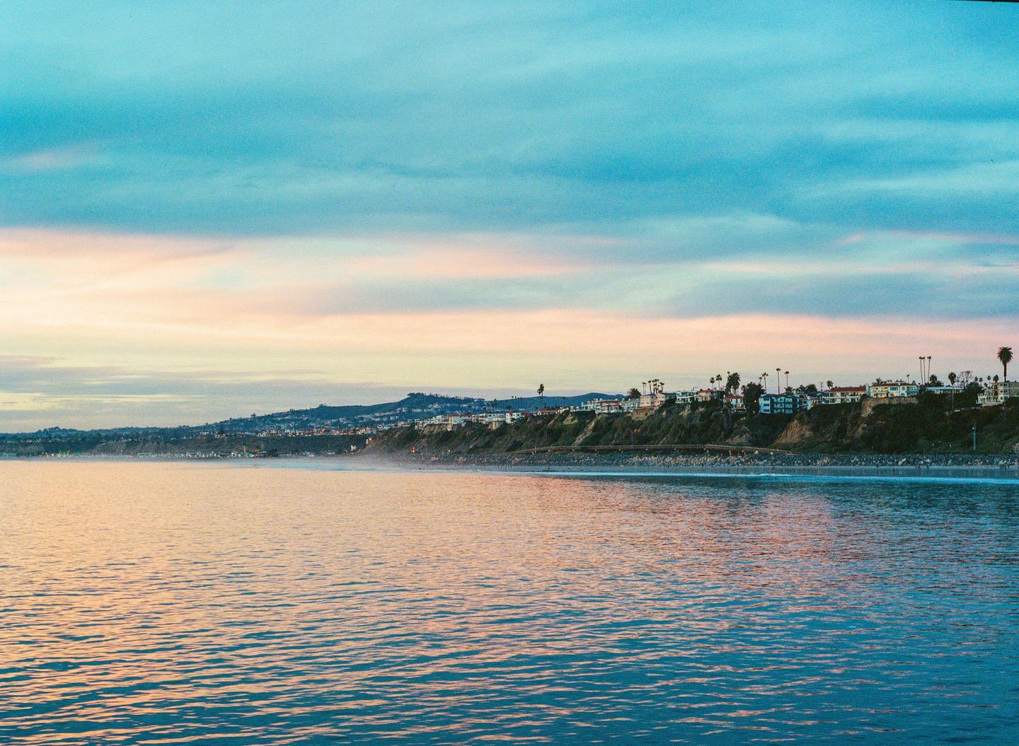 A coastal town with palm trees sits on a cliff overlooking the ocean under a colorful sky.