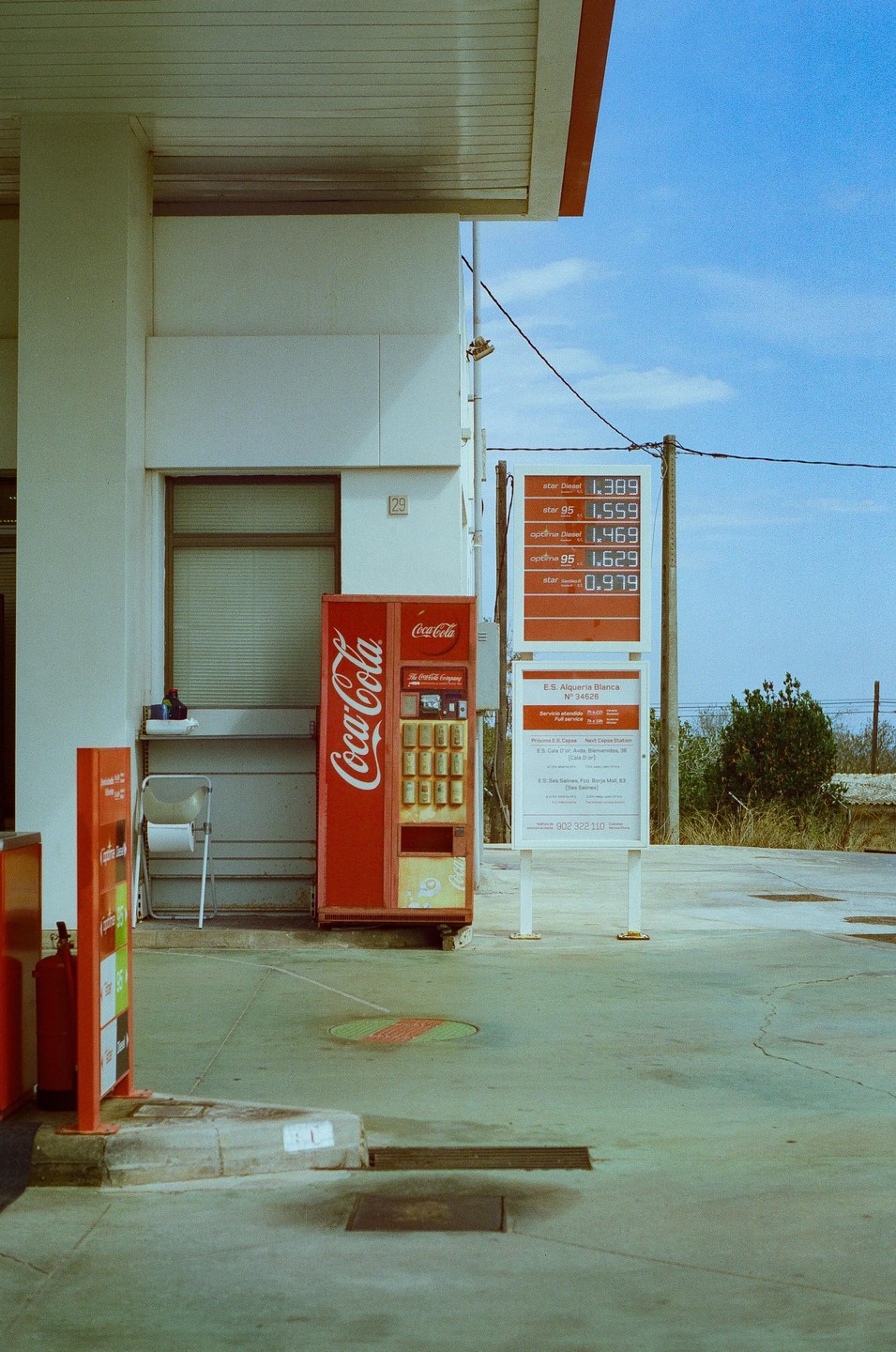 A red Coca-Cola vending machine stands next to a gas station building with a price sign under a blue sky.