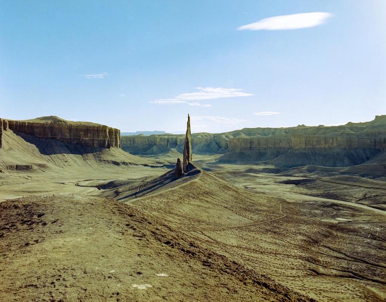 A tall, thin rock formation stands in a vast desert landscape under a blue sky.