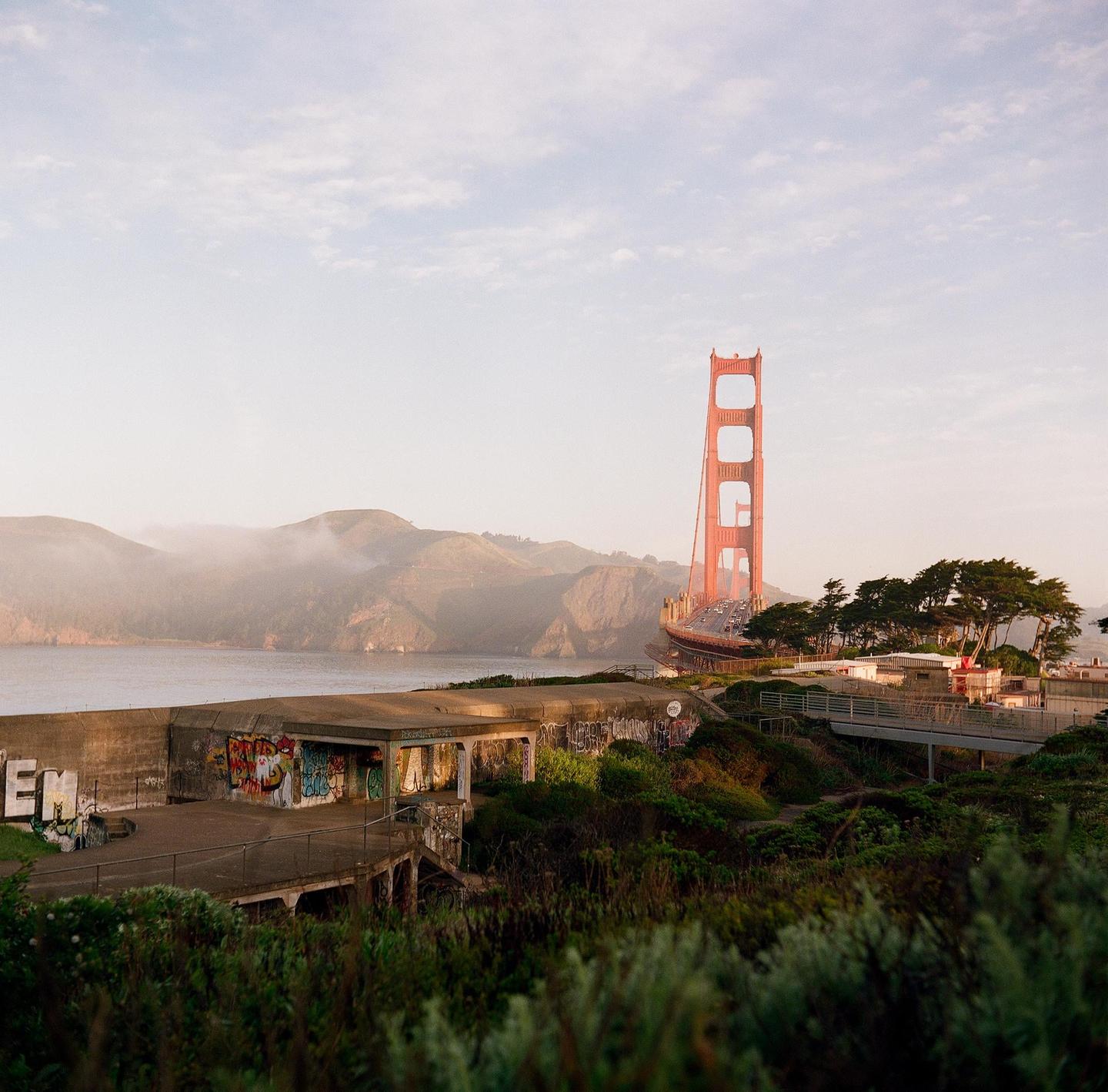 The Golden Gate Bridge is visible in the distance behind a concrete structure covered in graffiti and green foliage.