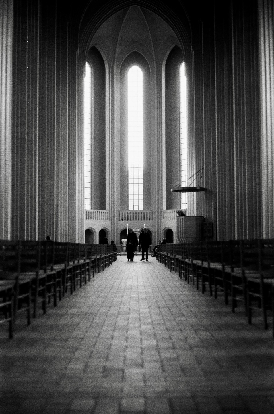 Two people walk down the aisle of a large church with tall, arched windows.