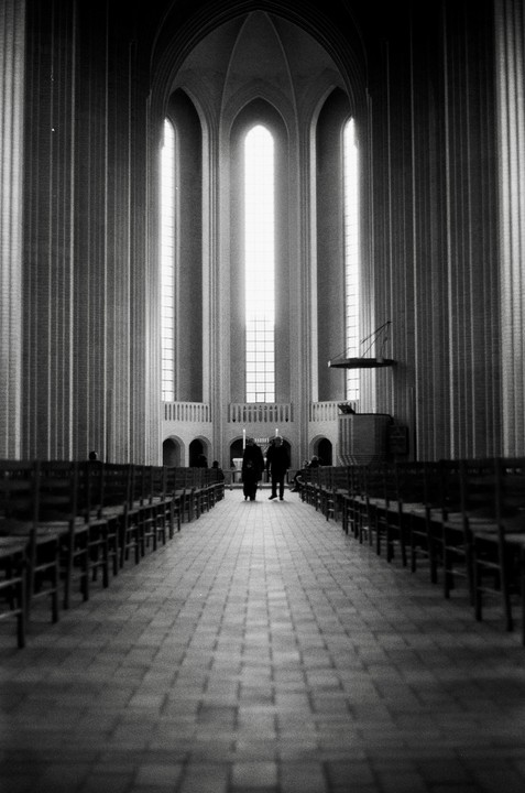 Two people walk down the aisle of a large church with tall, arched windows.