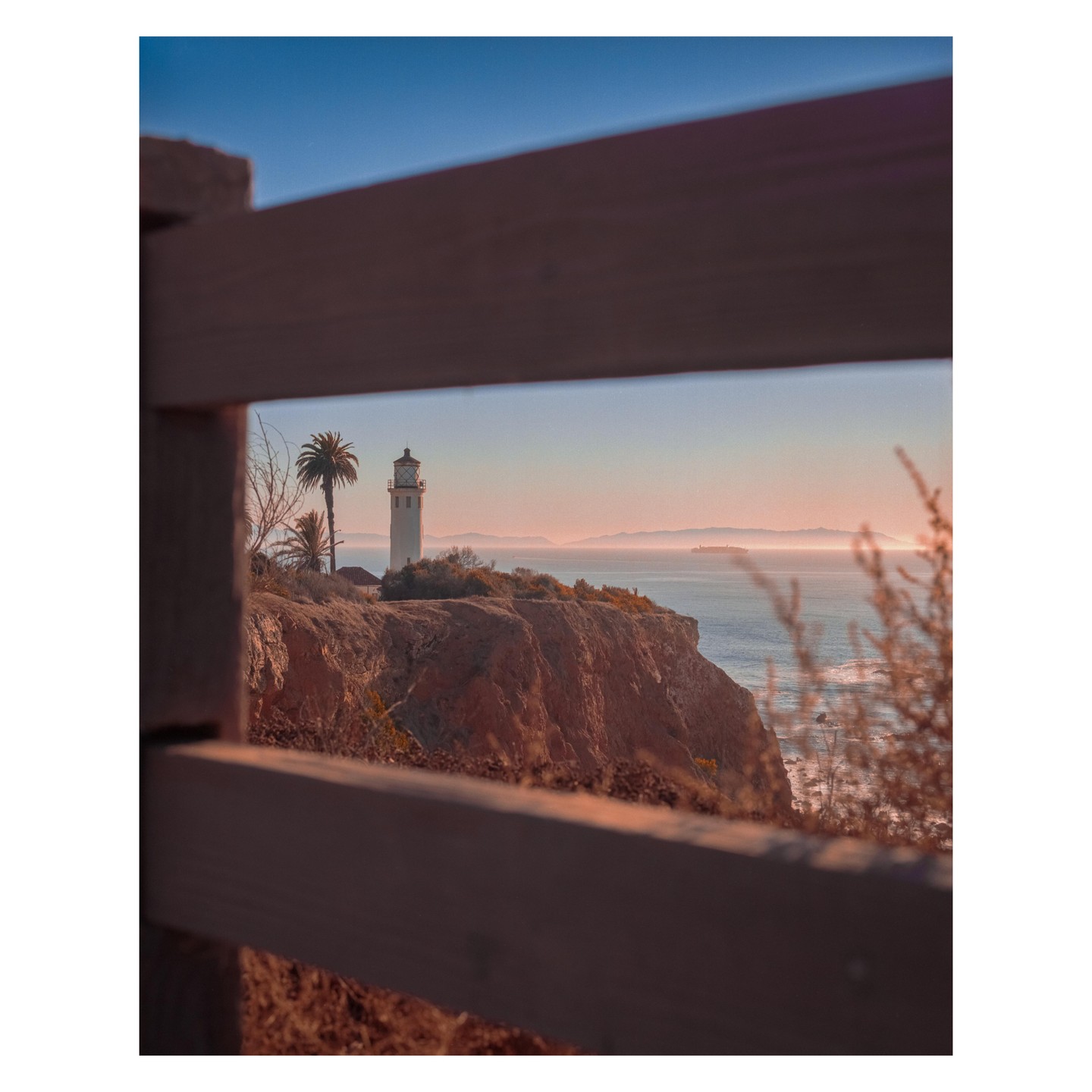 A lighthouse stands on a cliff overlooking the ocean at sunset, framed by a wooden fence.