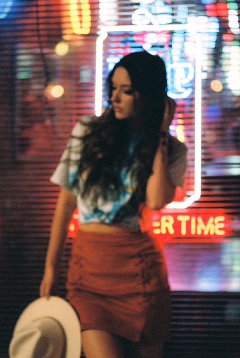 A woman with long hair holds a hat while standing in front of neon signs at night.
