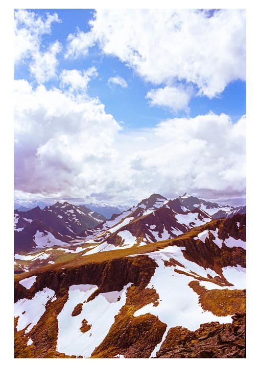 A vast mountain range with snow-capped peaks and patches of green and brown terrain stretches under a partly cloudy sky.