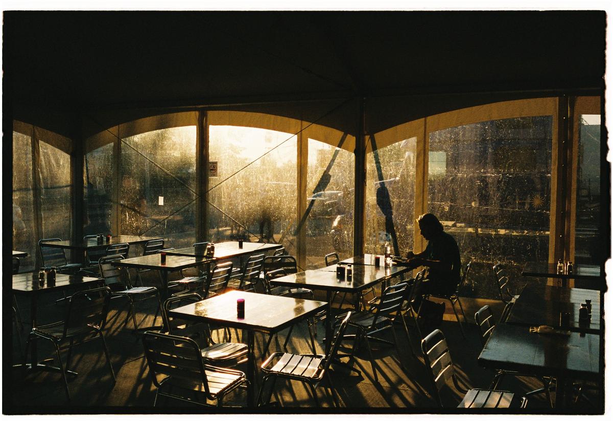 A person sits alone at a table in a restaurant with sunlight streaming through large windows.