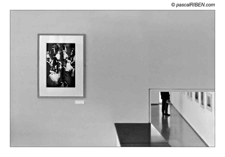 A framed black and white photograph of dancers hangs on a wall next to a cutout revealing a person standing in a hallway lined with framed pictures.