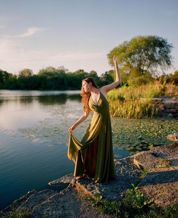 A woman in a green dress stands on rocks next to a lake with lily pads.