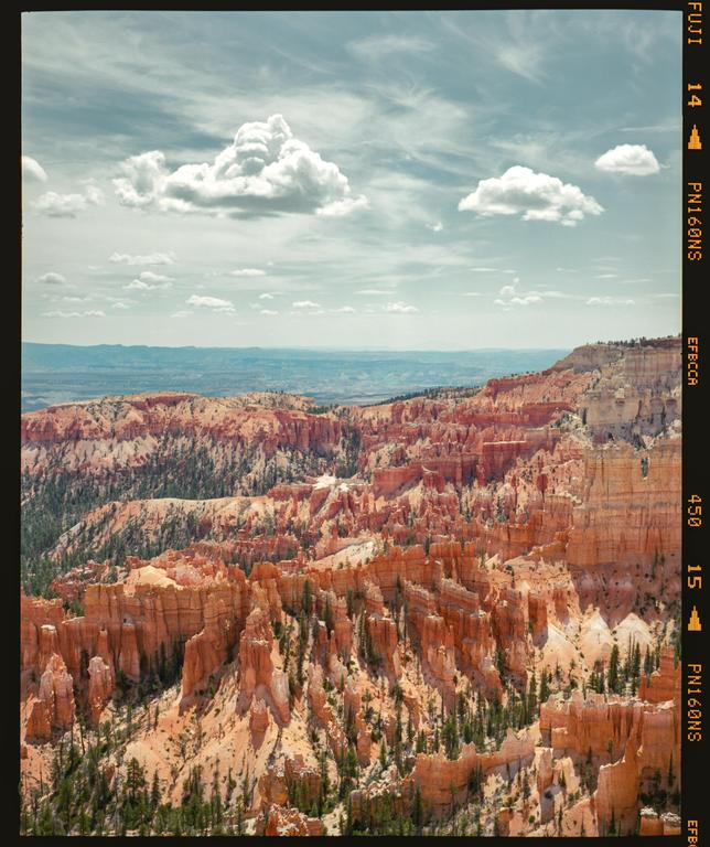 A wide view shows the orange hoodoos and green trees of Bryce Canyon National Park under a cloudy sky.