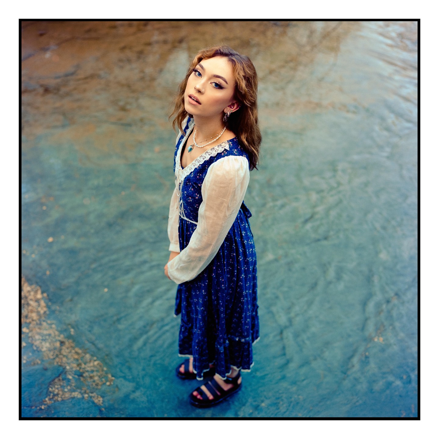 A young woman in a blue dress stands in shallow water, looking up at the viewer.
