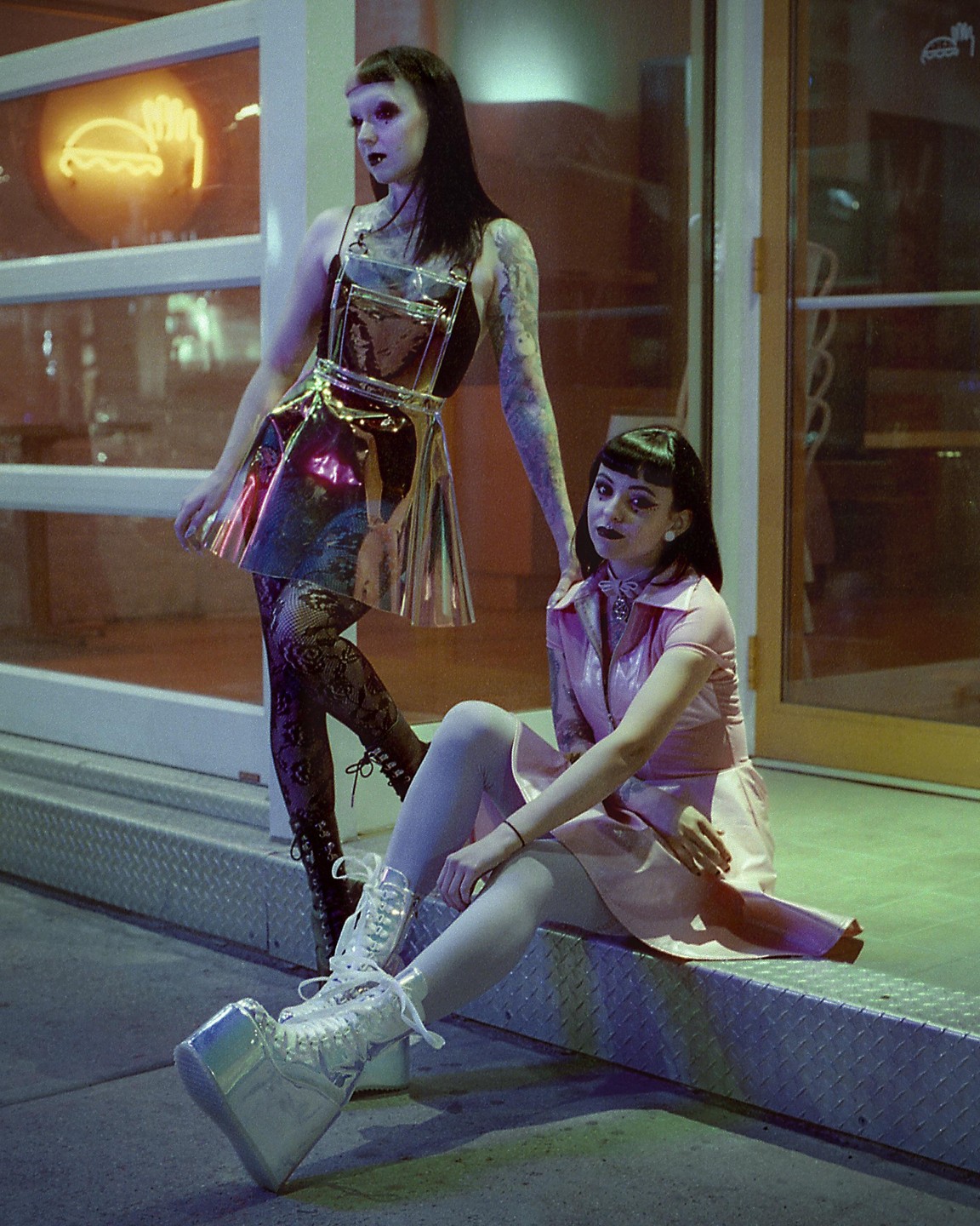 Two women pose at night in front of a building with a neon sign.