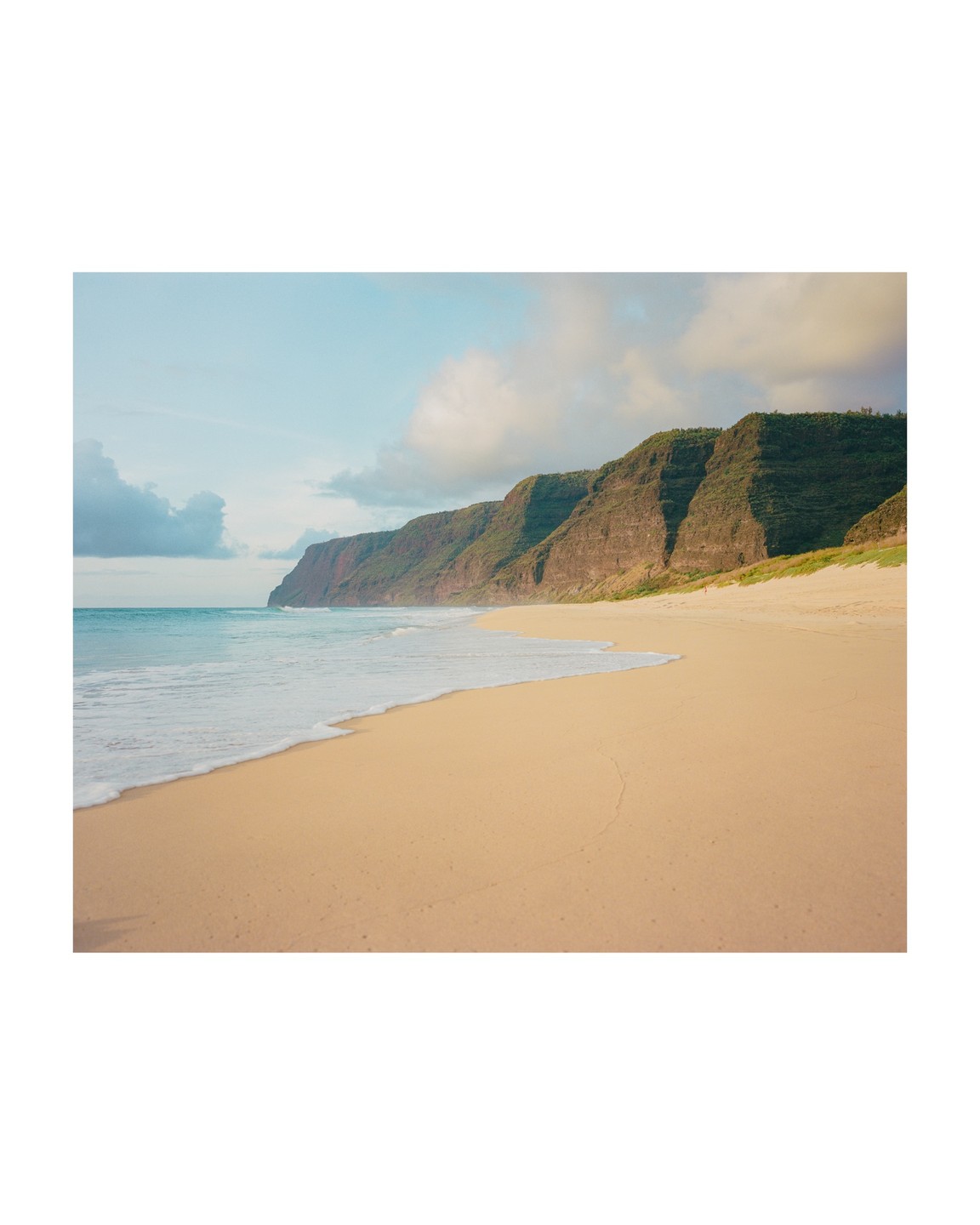 Waves wash onto a sandy beach backed by a line of steep, green cliffs under a partly cloudy sky.