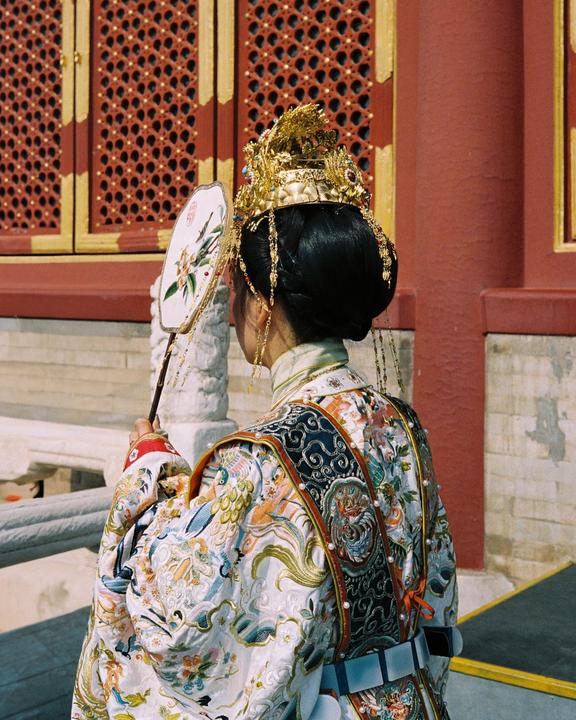 A woman in traditional attire with an elaborate golden headdress and a decorative fan stands with her back to the viewer.