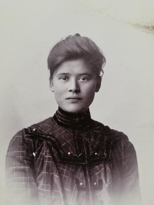 A woman with her hair up and wearing a plaid blouse with a high collar poses for a portrait.