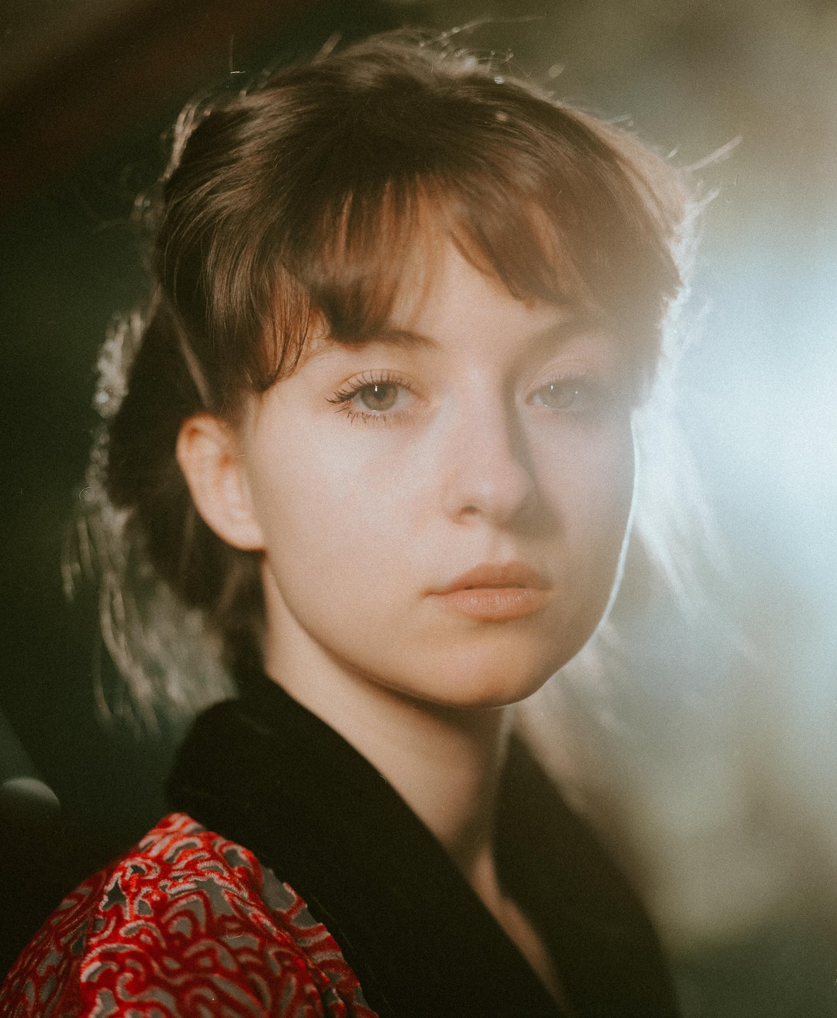 A woman with bangs and a patterned red and black garment looks directly at the viewer.