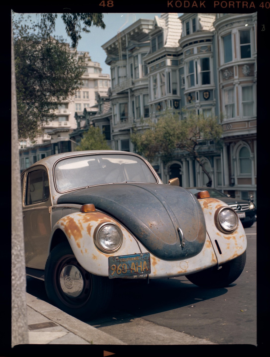 A rusty Volkswagen Beetle is parked on a street in front of ornate Victorian houses.