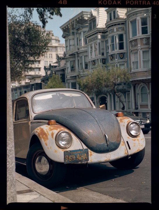 A rusty Volkswagen Beetle is parked on a street in front of ornate Victorian houses.