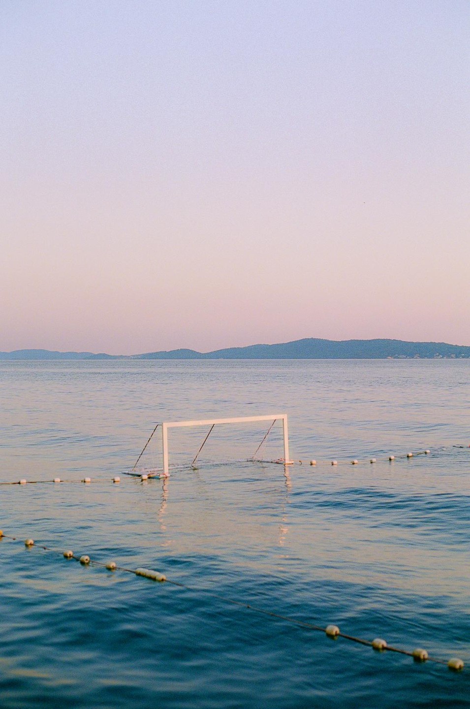 A white goal post stands in the water with a line of buoys stretching across the foreground and mountains in the distance under a pastel sky.