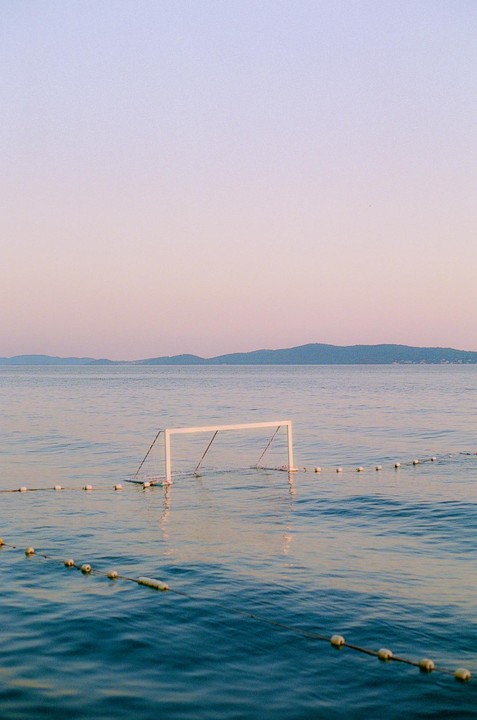 A white goal post stands in the water with a line of buoys stretching across the foreground and mountains in the distance under a pastel sky.