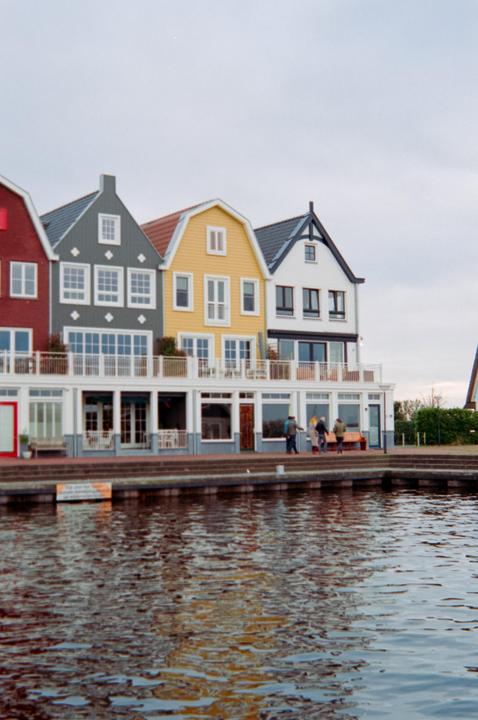 A row of colorful houses with balconies overlooks a body of water where people are walking along the shore.