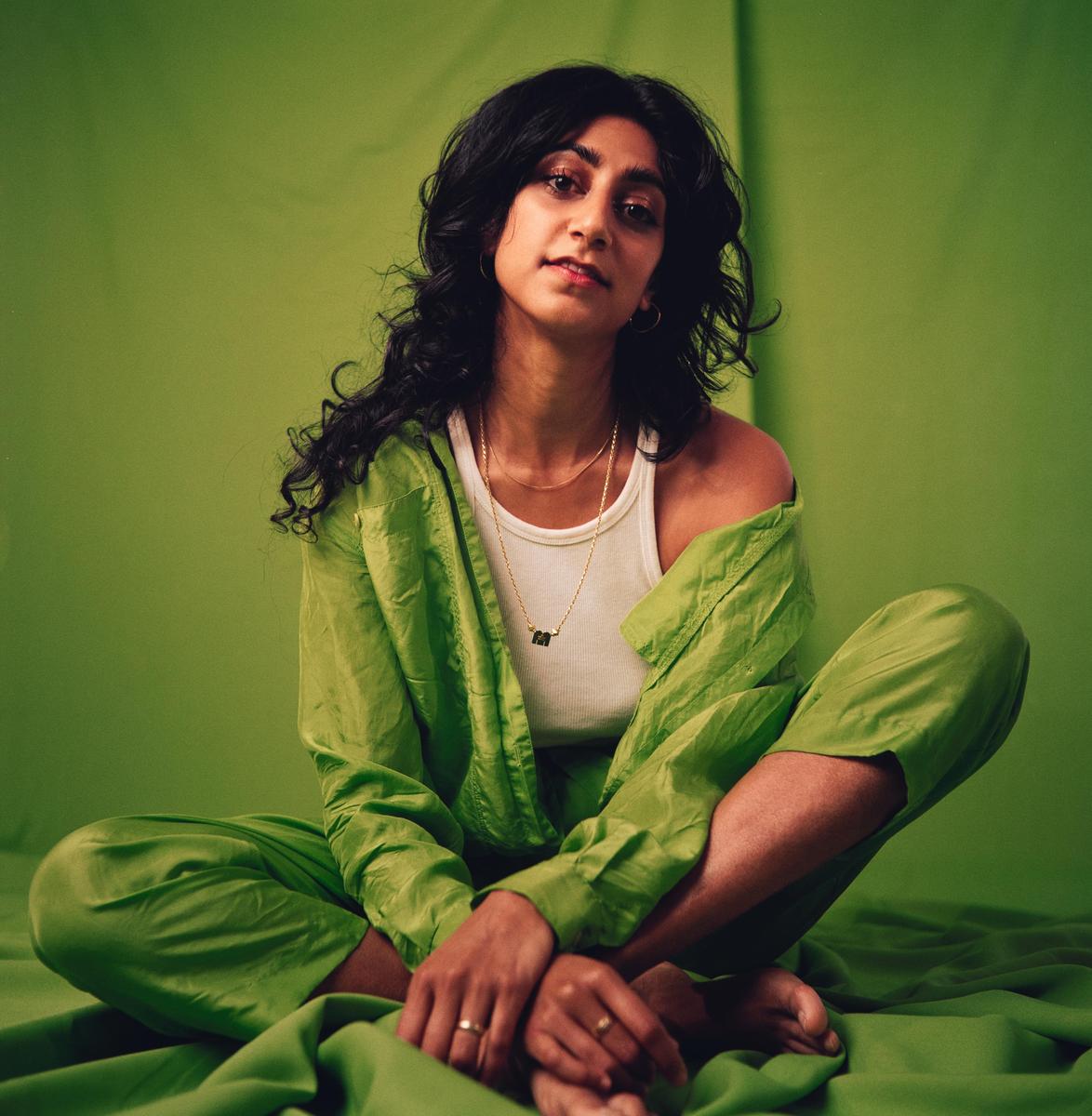 A woman with dark, curly hair sits cross-legged on a green sheet, wearing a white tank top and a green shirt.